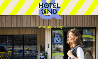 A modern hotel with an attractive facade of wood and colorful stripes. In the foreground stands a smiling woman enjoying the relaxed atmosphere.