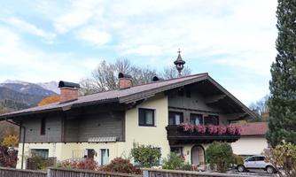 A traditional house in a rural setting with wooden cladding and a balcony full of flowers. In the background, mountains and a clear sky are visible.