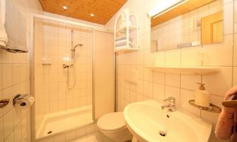 A modern bathroom with a shower, sink, and toilet. The walls are light-tiled and there are wooden paneling on the ceiling.