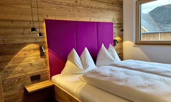 A modern bedroom with two single beds and a striking purple headboard wall. The room features wooden elements and offers daylight through a window.