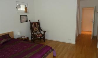 A bright bedroom with a large bed and a chair. The floor is made of wood and the walls are kept simple.