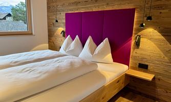 A modern bedroom with a double bed and two large white pillows. The wooden paneling and the purple wall give the room a cozy atmosphere.