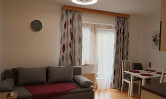 A cozy living-dining room with a gray sofa and a table for four people. Large windows with curtains let in plenty of light.