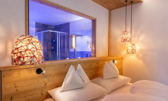 A modern bedroom with wooden details and soft lighting. In the background, a bathroom with a glass wall is visible.
