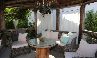 A cozy outdoor seating area with rattan furniture and a wooden table. Soft cushions and a beautiful chandelier complete the relaxed atmosphere.