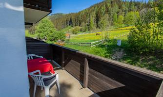 A balcony with a table and chairs overlooks a green landscape. In the background, trees and meadows can be seen.