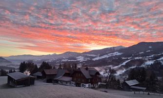 A picturesque winter landscape with a farmhouse and snow-covered mountains. The sky is illuminated in vibrant colors at sunrise.