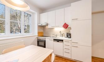 A modern kitchen with white cabinets and a wooden floor. There are windows that let in plenty of daylight.