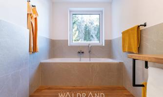 A modern bathroom with a large bathtub and bright wood flooring. The walls are in neutral tones, and orange towels provide a splash of color.