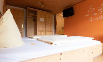 A cozy room with a wooden bed and soft cushions. The walls are painted in warm colors, featuring a TV corner and a wardrobe.