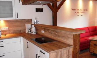 A modern kitchen with wooden elements and a cooking island. In the background, a cozy living room with a red sofa can be seen.