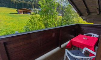 A balcony with a red table and chairs overlooks a green meadow and wooden chalets. The surroundings are surrounded by trees and convey a serene rural atmosphere.