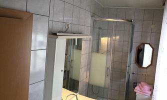 A bright bathroom with a shower and a sink. The walls are tiled and there is a mirror as well as a wooden shelf.