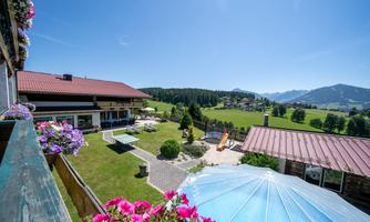 A picturesque view of the mountains and the green landscape with colorful flowers. In the foreground, there is a well-kept garden and a terrace with a sun umbrella.