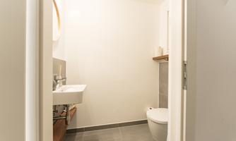 A modern bathroom with a simple sink and a toilet. The walls are bright and the floor is covered with grey tiles.