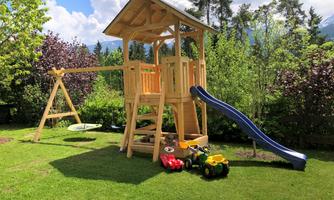 A playground in the garden with a wooden play castle, a slide, and a swing. Toys like a red and a yellow car are visible in the foreground.