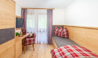 A cozy room with wooden interiors and a single bed. The seating area is decorated with red-checked cushions and a window provides plenty of light.