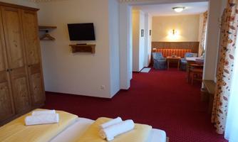 A cozy room with two single beds and a red carpet. In the background, a living area with a table and chairs, as well as a television, is visible.