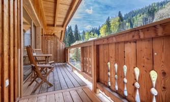A cozy wooden balcony with two chairs. In the background, green trees and a clear sky can be seen.