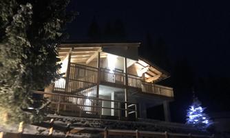 A modern wooden house at night, surrounded by trees. A lit Christmas tree stands on the premises.