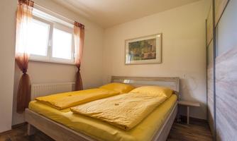 A cozy bedroom with a double bed and yellow comforters. The walls are light, and there are large windows with curtains.