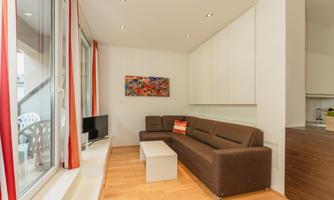 A modern living room with a large window and bright walls. The furnishing consists of a brown sofa, a white table, and a television.