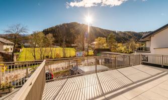 A sunny terrace with a view of the mountains. The sky is clear and the landscape is green and autumnal.