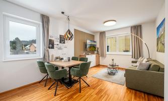 A modern living room with a dining area and a sofa. Large windows provide plenty of daylight and a cozy atmosphere.