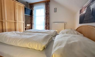 A cozy bedroom with a double bed and white bed covers. A window provides plenty of daylight, and the room is simply furnished.