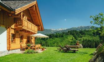 A charming wooden house in a green landscape with mountains in the background. The garden offers a cozy seating area with an umbrella.