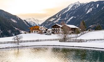 A picturesque winter landscape with snow-covered mountains in the background. In the foreground, there is a tranquil pond and traditional farmhouses surrounded by snowy meadows.