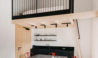 A modern apartment with a loft area above and a dining area below. Red chairs and a black kitchen table create a stylish contrast.