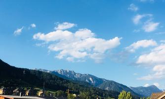 A picturesque mountain landscape under a clear blue sky with some clouds. In the foreground, houses and trees are visible.