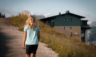 A girl with long hair walks along a path towards a building in the alpine landscape. The surroundings are green and mountainous, with some mist in the distance.