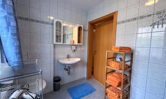 A modern bathroom with white tiles and a sink. Towels in various colors are stacked on a shelf.