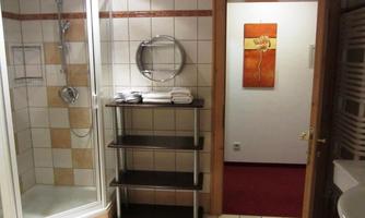 A bathroom with a shower and a shelf for towels. The adjoining room has a red carpet and a decorative picture on the wall.