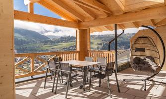A sunny terrace with a wooden structure and a view of the mountains. The area is equipped with a table, chairs, and a hammock.