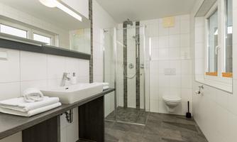 A modern bathroom with a glass shower and bright tiles. The sink area is equipped with fresh towels and an elegant shelf.