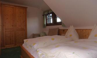 A cozy bedroom with a double bed and wooden furniture. There is a wardrobe and a window overlooking the outside.