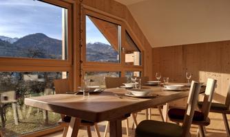 A modern dining room with wooden interiors and large windows. The view overlooks a picturesque mountain landscape.
