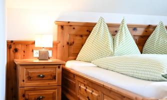 A cozy bedroom with a wooden bed and checkered pillows. Next to the bed is a nightstand with a small lamp.