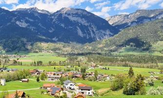A picturesque landscape with green meadows and charming villages. In the background, high mountains rise under a blue sky.