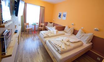 A cozy hotel room with two beds, on which decorative towels are placed. The walls are painted orange, and there are windows with curtains as well as a television.