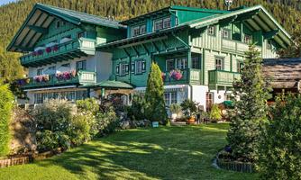 A beautiful, traditional house with a green facade and colorful flower balconies. The garden is well-maintained and surrounded by trees, with hills visible in the background.