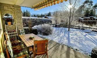 A cozy terrace with a wooden table and chairs, surrounded by snow-covered trees. The sun shines over the snowy landscape.
