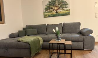 A cozy living room with a gray sofa and a small table. A picture of a tree hangs on the wall and a green blanket lies on the sofa.