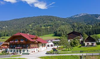 An idyllic farmhouse in a picturesque landscape. In the background, there are green hills and trees as well as a clear blue sky.