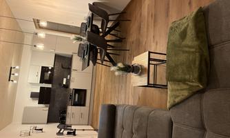 A modern kitchen with a dining table and chairs in a cozy living area. The room has wooden flooring and warm lighting.