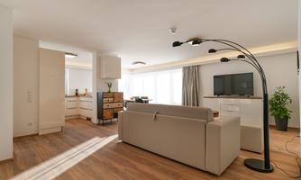 A modern living room with a beige sofa and large windows. The room is brightly lit and has an open connection to the kitchen.