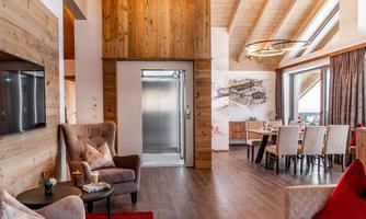 A modern kitchen-living room with wooden decor and large windows. An elevator is visible, and the space is stylishly and invitingly furnished.
